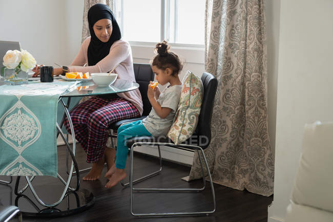 Vista lateral de la raza mixta Madre usando hijab hablando con su hija mientras come fruta naranja mientras está sentada en la mesa en casa - foto de stock