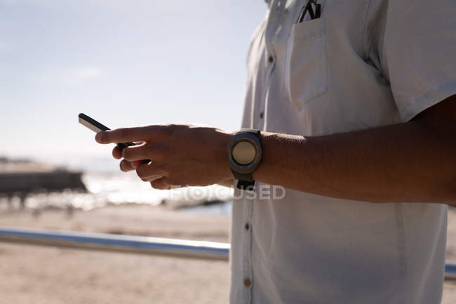 Sección media del hombre de raza mixta usando teléfono móvil en la playa bajo el sol - foto de stock