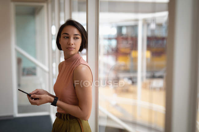 Vista lateral de uma empresária asiática atenciosa segurando seu telefone celular enquanto olha pela janela do escritório — Fotografia de Stock