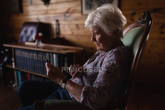 Vista lateral de uma mulher idosa ativa usando um telefone celular enquanto sentada em uma poltrona em casa — Fotografia de Stock
