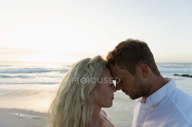 Vista lateral de la joven pareja de amor avergonzándose mientras están de pie en la playa. Están disfrutando de sus vacaciones - foto de stock