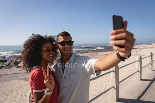 Вид спереди на афро-американскую пару, стоящую и говорящую селфи, улыбающуюся на пляже в солнечный день — стоковое фото