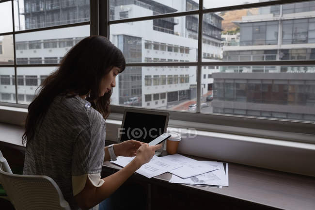 Side view of business woman using mobile phone in office at desk — стоковое фото