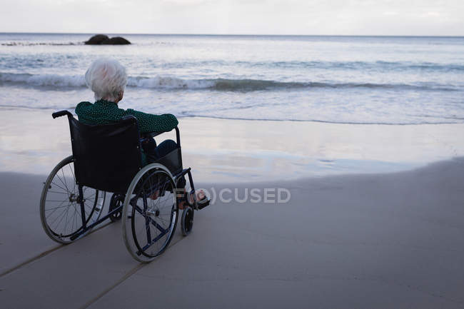 Вид сзади на активную пожилую женщину-инвалида, смотрящую на море, сидя на инвалидном кресле на пляже — стоковое фото