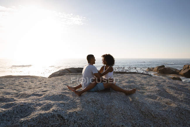 Vista lateral de una pareja afroamericana en un ambiente romántico sentada en la roca cerca del mar y mirándose unos a otros - foto de stock