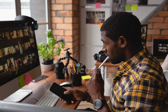 Vista lateral del hombre de negocios reflexivo afroamericano que trabaja sobre la computadora sosteniendo el teléfono móvil en la mano en la oficina - foto de stock