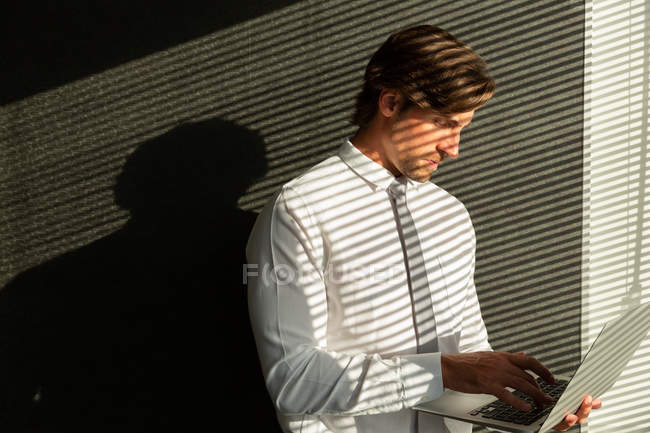 Vue de face de beau jeune cadre masculin travaillant sur ordinateur portable tout en se tenant près de la fenêtre dans un bureau moderne — Photo de stock