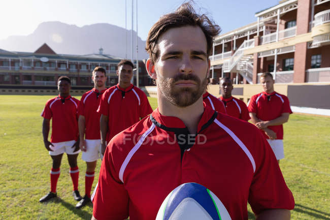 Vista frontal de uma equipe de jogadores de rugby multi étnicos masculinos de pé com uma bola de rugby no chão de rugby em um dia ensolarado — Fotografia de Stock