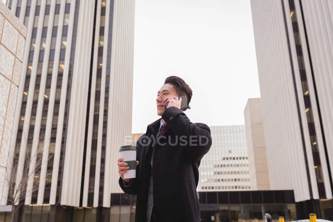 Front view of young Asian businessman with coffee cup talking on mobile phone in city surrounded by tall business buildings — Stock Photo