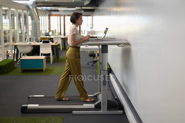 Vue latérale d'une femme d'affaires asiatique travaillant sur son ordinateur portable tout en faisant de l'exercice sur tapis roulant dans le bureau — Photo de stock