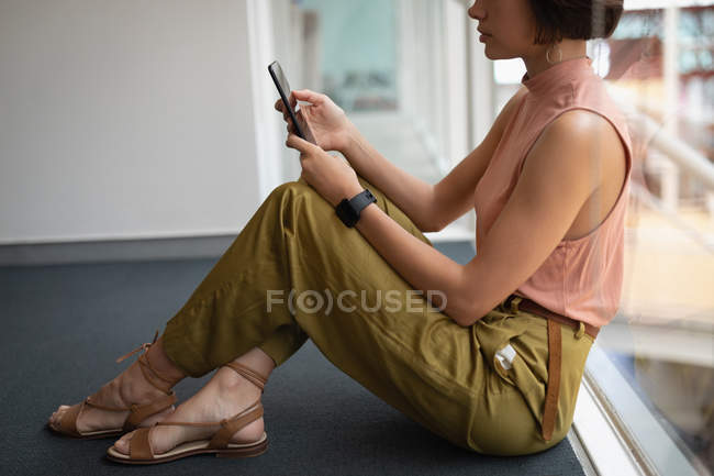 Vista laterale di una donna d'affari asiatica che utilizza il suo telefono cellulare mentre si appoggia a una finestra in ufficio — Foto stock