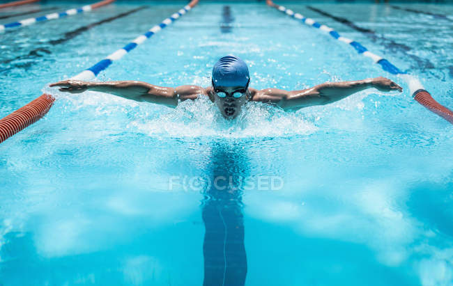 Front view of young Caucasian male swimmer swimming butterfly stroke in outdoor swimming pool on bright sunny day — Stock Photo