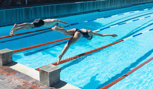 Vista lateral de nadadores caucasianos masculinos e femininos pulando na água ao mesmo tempo na piscina ao sol — Fotografia de Stock