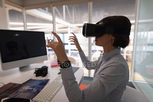 Vista lateral de una mujer de negocios asiática usando un auricular de realidad virtual en el escritorio en la oficina - foto de stock