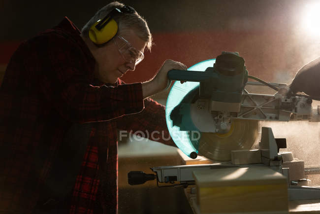 Vista lateral del carpintero con auriculares cortando una pieza de madera con una sierra mecánica en el taller - foto de stock