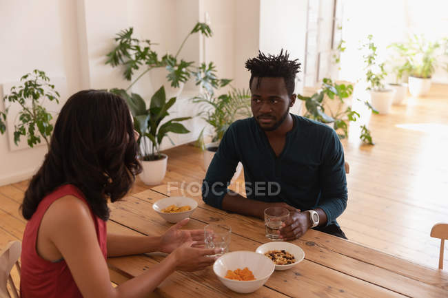 Вид спереду багатонаціональна пара сидить і взаємодіє один з одним вдома, маючи закуски навколо столу — стокове фото