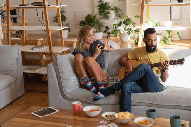Front view of Woman using mobile phone while mixed race man playing guitar in living room at home — Stock Photo