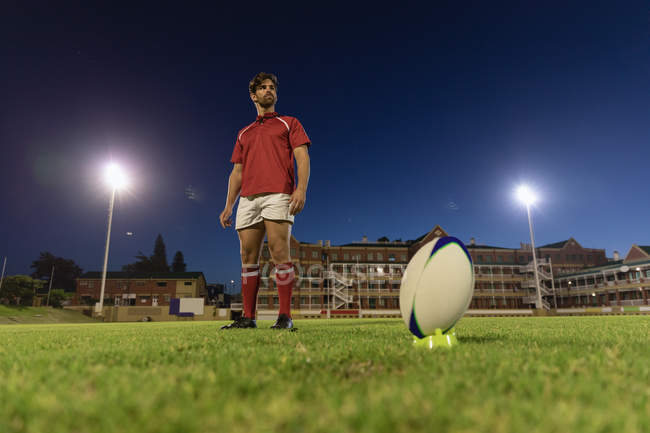 Vista de ángulo bajo de un jugador de rugby masculino de pie y esperando para patear la pelota de rugby en el estadio en la noche - foto de stock