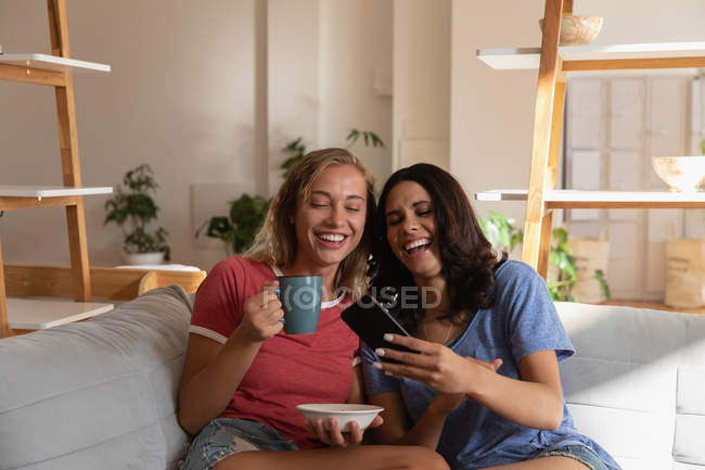 Vista frontal de las amigas divirtiéndose mientras usan el teléfono móvil en la sala de estar en casa - foto de stock