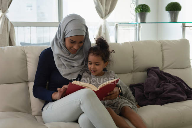 Вид спереди матери со смешанным расовым происхождением в хиджабе и дочери, сидящей и читающей книгу в гостиной дома — стоковое фото