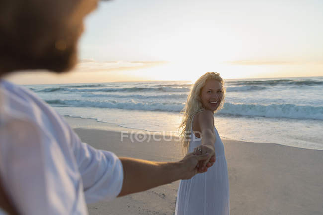 Vista lateral do jovem casal de amor de mão dada enquanto está de pé na praia em um dia ensolarado. Eles estão desfrutando suas férias — Fotografia de Stock