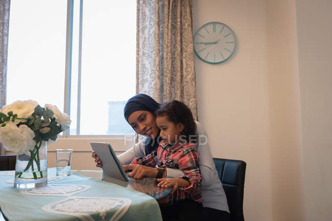 Vue latérale de mère métissée portant hijab et fille utilisant une tablette numérique à la maison sur une chaise autour d'une table dans le salon — Photo de stock