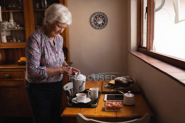Вид сбоку активной пожилой женщины, наливающей кофе в чашку за обеденным столом на кухне дома — стоковое фото
