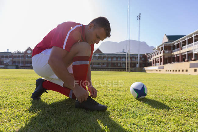 Vista lateral de um jogador de rugby caucasiano macho amarrando seus atacadores no chão de rugby ao sol — Fotografia de Stock
