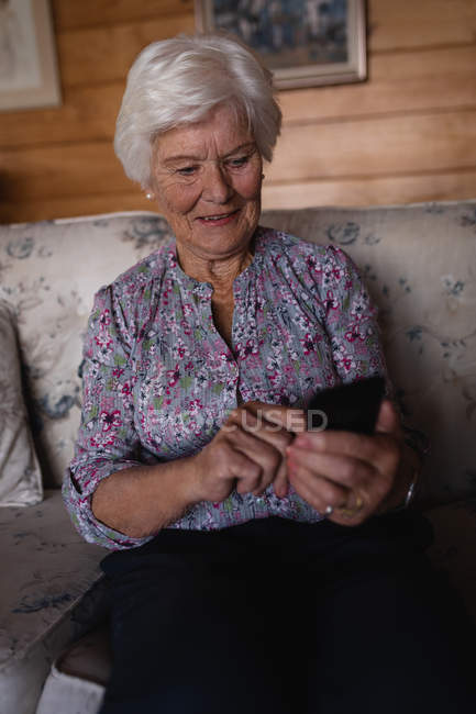 Вид спереди счастливой активной пожилой женщины, которая пользуется мобильным телефоном, сидя дома на диване — стоковое фото