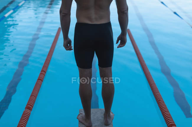 Section basse arrière d'un nageur mâle debout sur le bloc de départ devant la piscine — Photo de stock