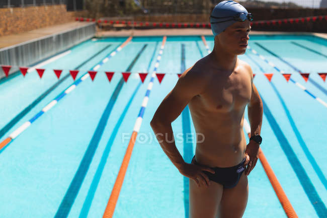 Vista frontal do jovem nadador caucasiano do sexo masculino de pé com as mãos no quadril enquanto veste roupa de banho e assista na piscina exterior no dia ensolarado — Fotografia de Stock
