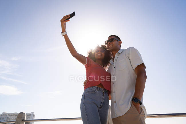 Baixo ângulo de visão do casal afro-americano de pé e falando selfie na praia ao sol — Fotografia de Stock