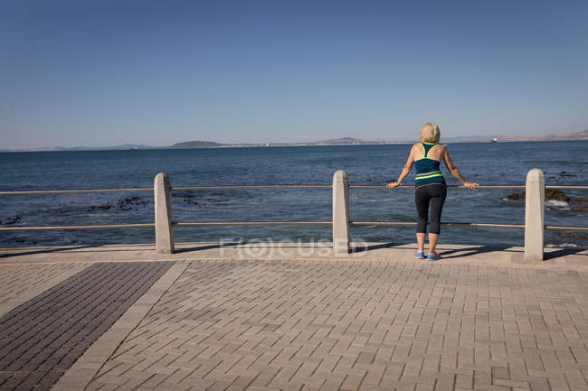 Visão traseira de uma mulher idosa ativa apoiando-se contra o trilho de guarda de um passeio em frente ao mar sob o sol — Fotografia de Stock