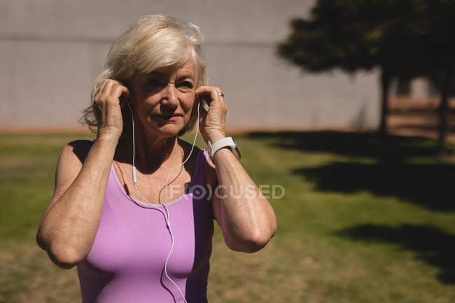 Vista frontale di una donna anziana attiva che indossa gli auricolari per ascoltare musica mentre si allena nel parco in una giornata di sole — Foto stock
