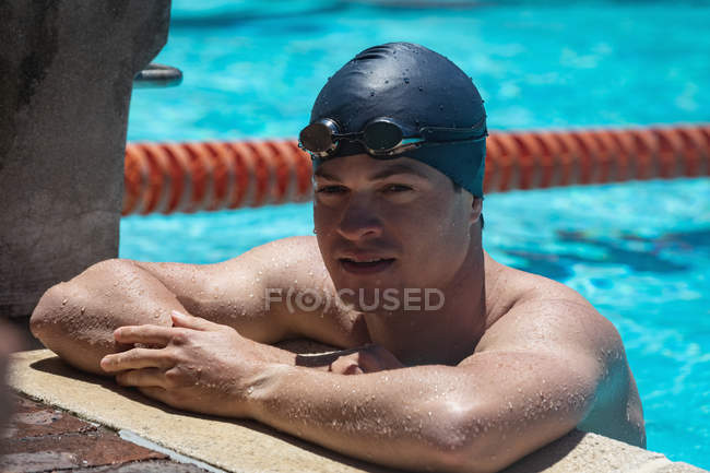 Close-up portrait of young Caucasian male swimmer standing at the edge of a pool and looking at camera in the sunshine — Stock Photo