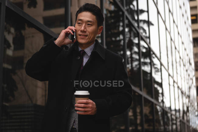 Vista frontal de un joven empresario asiático bien vestido con taza de café desechable hablando por teléfono móvil en la ciudad - foto de stock