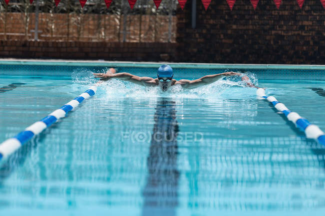 Vista frontal de un joven nadador caucásico trabajando duro mientras nade un golpe de mariposa en la piscina al aire libre bajo el sol - foto de stock