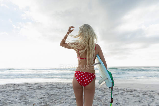Vista trasera de la rubia surfista femenina con una tabla de surf de pie en una playa en un día soleado. Ella camina hacia el océano - foto de stock