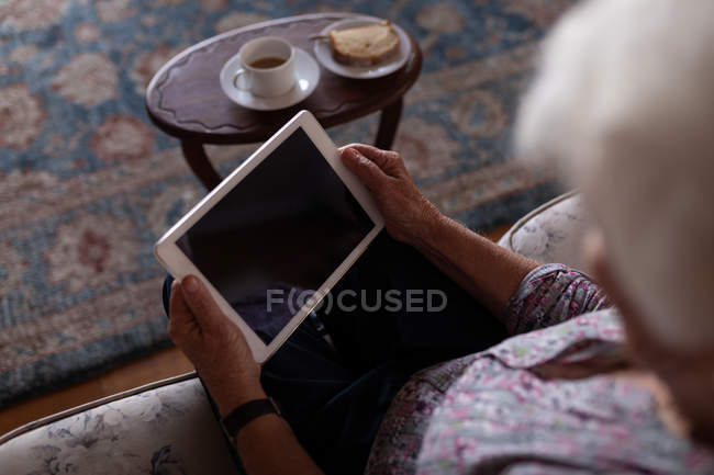 Vista de ángulo alto de una mujer mayor activa usando una tableta digital mientras está sentada en el sofá en casa para tomar el té - foto de stock