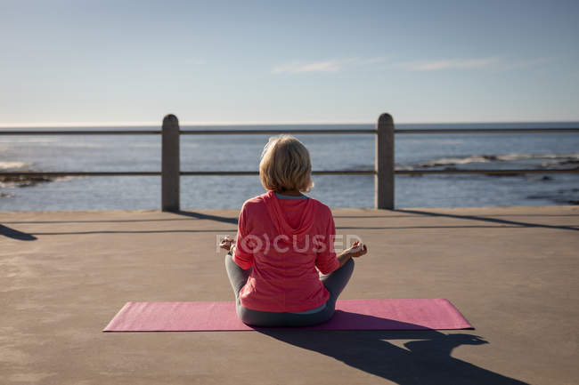 Vista posteriore di una donna anziana attiva che esegue yoga su una mappa di fitness su una passeggiata di fronte al mare — Foto stock