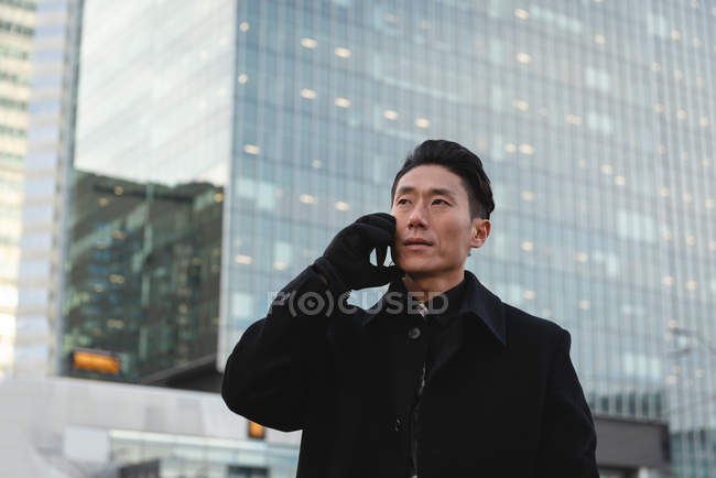 Низкий угол обзора молодой азиатский бизнесмен разговаривает по мобильному телефону, стоя на улице со зданием позади него в городе — стоковое фото
