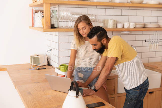 Vista lateral de la pareja multiétnica de pie y trabajando sobre el ordenador portátil en el hogar en la sala de cocina - foto de stock