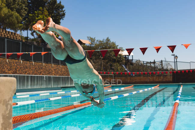 Low angle view of young Caucasian male swimmer diving into water of empty outdoor swimming pool on sunny day — Stock Photo