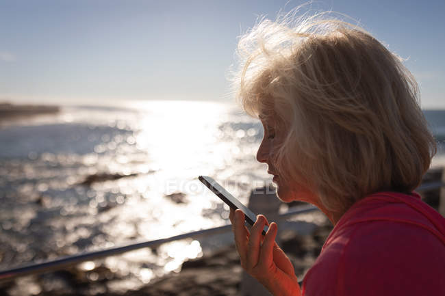 Крупный план активной пожилой женщины, разговаривающей по мобильному телефону на набережной перед морем в лучах солнца — стоковое фото