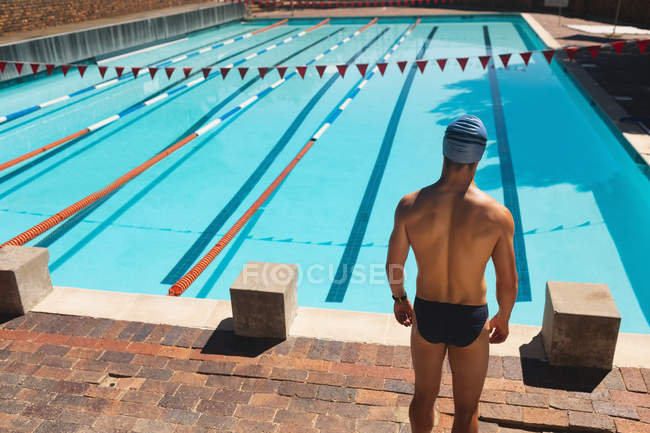 Rear view of young Caucasian male swimmer standing at the edge of outdoor swimming pool on sunny day — Stock Photo
