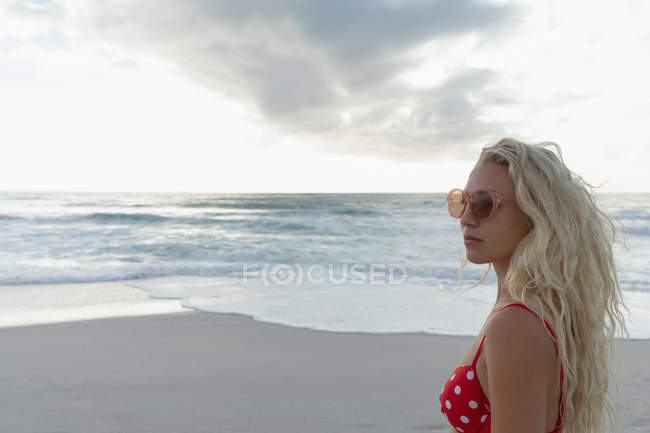 Vista lateral da bela mulher loira em pé na praia em um dia ensolarado. Ela usa óculos solares. — Fotografia de Stock