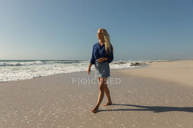 Vista frontal de la mujer rubia de pie en la playa en un día soleado. Ella está caminando. - foto de stock