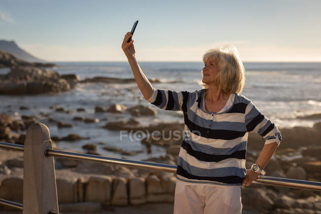 Vista frontal de una mujer mayor activa tomando una selfie con su teléfono móvil mientras se apoya contra una barandilla en un paseo marítimo en la noche para la puesta del sol - foto de stock