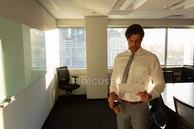 Vista frontal do jovem executivo masculino com xícara de documentos de leitura de café em um escritório moderno — Fotografia de Stock