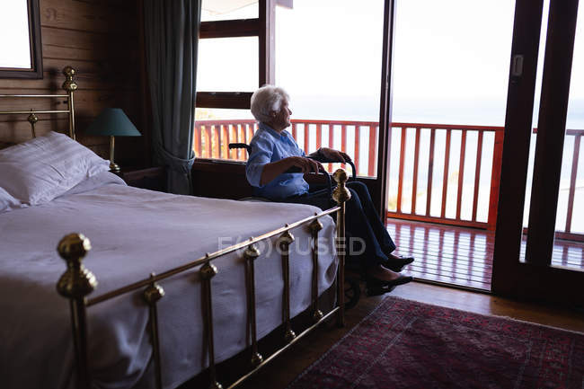 Side view of a disabled active senior woman sitting on a wheelchair and looking through the window in bedroom at home — Stock Photo